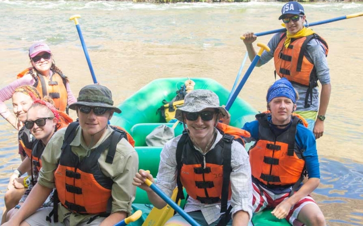 A group of people wearing life jackets sit in or stand near a raft floating in shallow water and smile at the camera